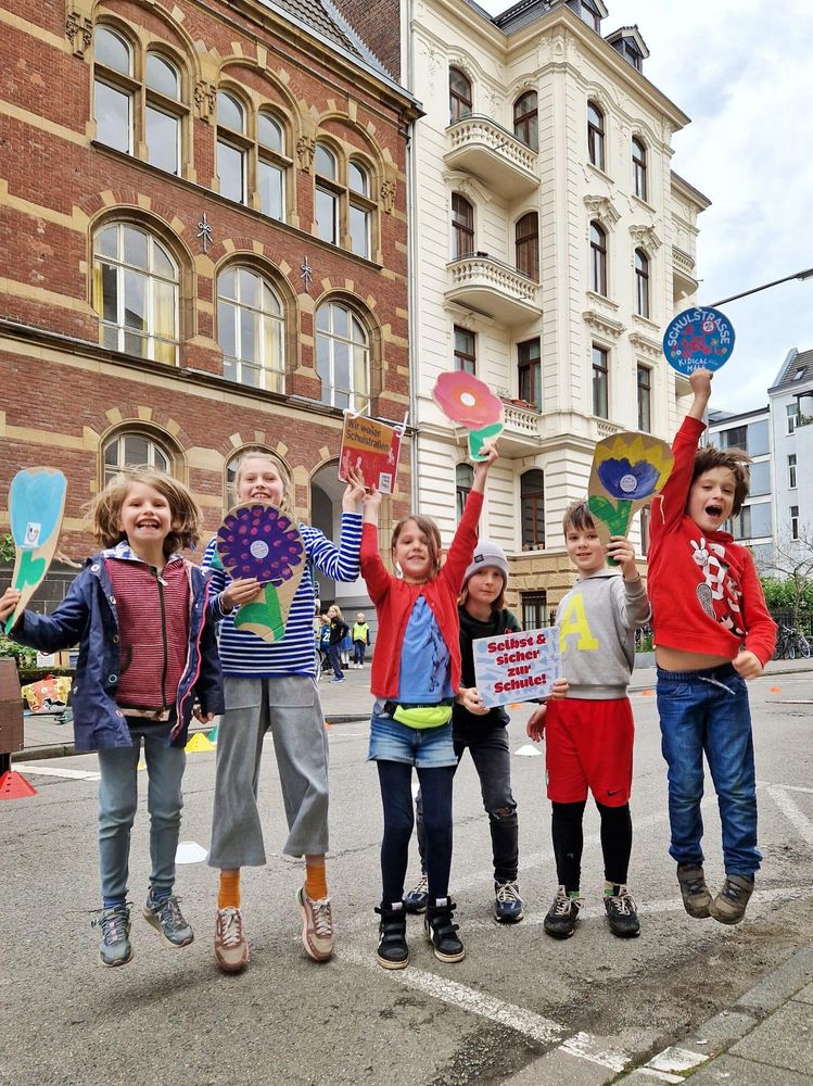 Sechs Kinder stehen vor einem Schulgebäude und springen lachend in die Luft. Sie halten bunte Schilder in Blumenform und Plakate hoch – eines trägt die Aufschrift: „Selbst & sicher zur Schule!“, ein anderes: „Wir wollen Schulstraßen“. Die Kinder wirken fröhlich, entschlossen und aktiv. Das Bild transportiert eine klare Botschaft: Kinder wollen sich sicher und selbstständig im Straßenverkehr bewegen – insbesondere auf dem Weg zur Schule.