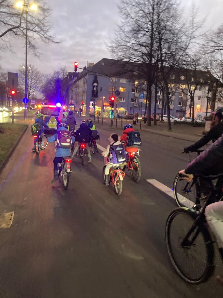 Eine Gruppe von Kindern und Erwachsenen auf Fahrrädern fährt bei Dämmerung auf einer städtischen Strasse. Die Kinder tragen Schulranzen und Helme, während ein Polizeiauto mit eingeschaltetem Blaulicht die Gruppe begleitet. Im Hintergrund sind Wohnhäuser, Bäume ohne Laub sowie eine Ampel zu sehen.  
