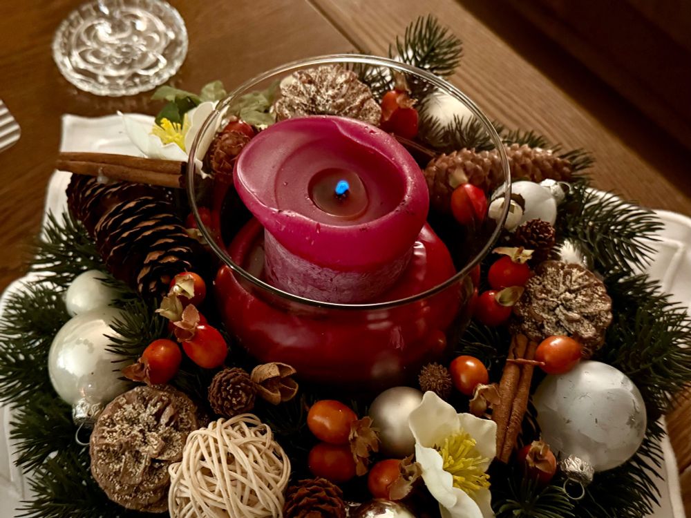 Ein größeres Weihnachtsgesteck mit einer großen, benutzten dunkelroten Kerze in einem Glas in der Mitte mit einem sehr mickrigen, bläulichen Flämmchen