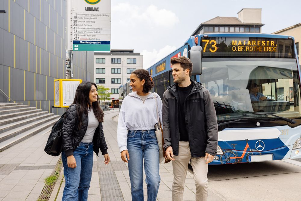 Drei Studierende der RWTH stehen stehen vor dem Hörsaalgebäude CARL vor einer Bushaltestelle, während neben ihnen ein Bus hält.