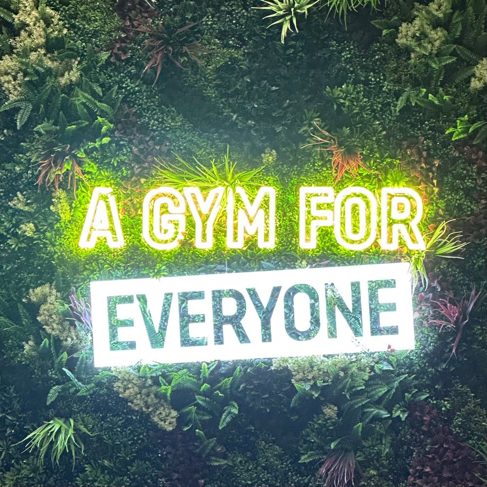 Lit letters of a sign, against a fake plant wall. “A gym for everyone”