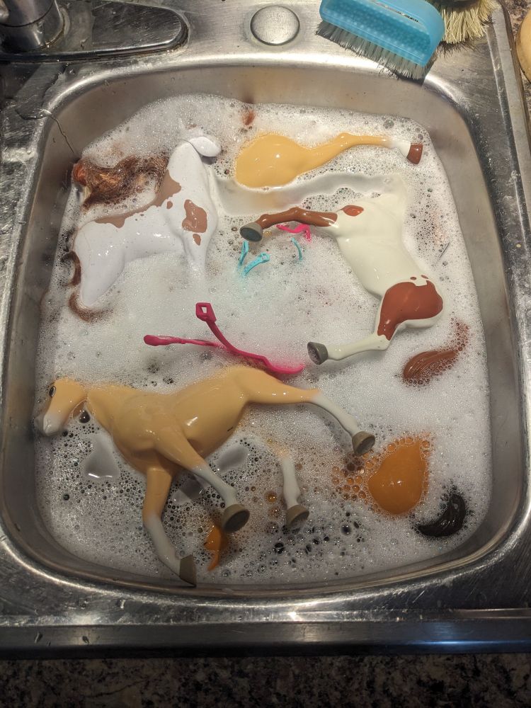 A photo of a kitchen sink full of soapy water and plastic toy horses.