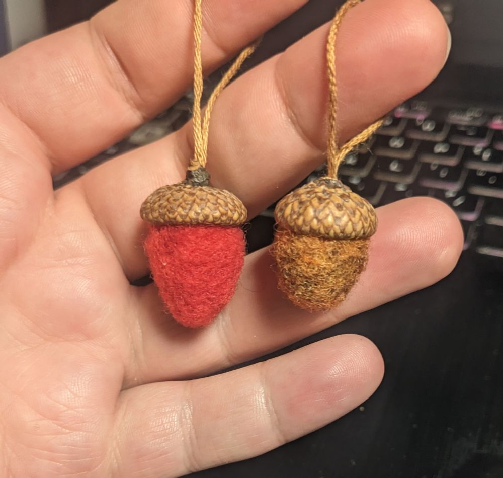 A photo of two needlefelted acorns with real acorn caps, on strings.
