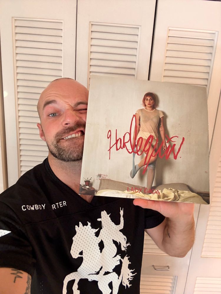 Image of a man wearing a t-shirt with a woman riding a horse. He holds the LP cover for Harlequin by Lady Gaga.