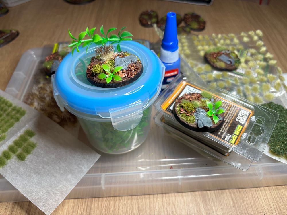Several containers of varied basing tufts with four forest themed bases on top of them and a bottle of superglue