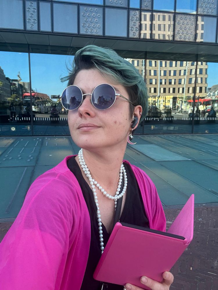 Blue haired woman using a bright pink long vest and holding a bright pink kindle