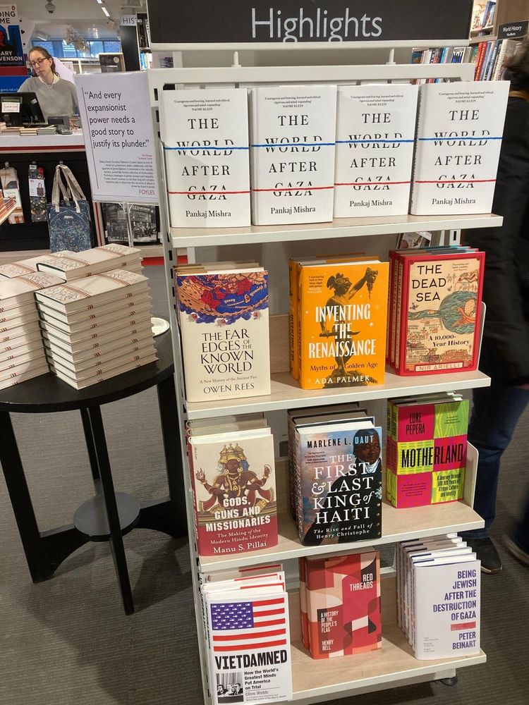 A set of book shelves with the black heading above saying 'Highlights'. Multicoloured book covers fill each shelf, three across. They are all history titles. In the background are a table with more white books stacked up and a shop till with a cashier wearing grey.