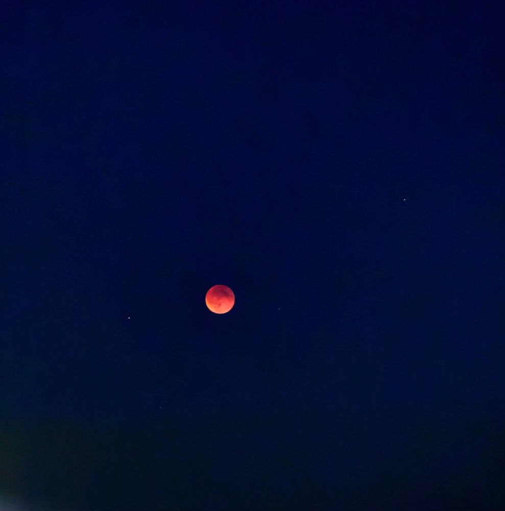 Das Bild zeigt einen dunklen Nachthimmel mit einem rötlich schimmernden Mond, umgeben von einigen Sternen.