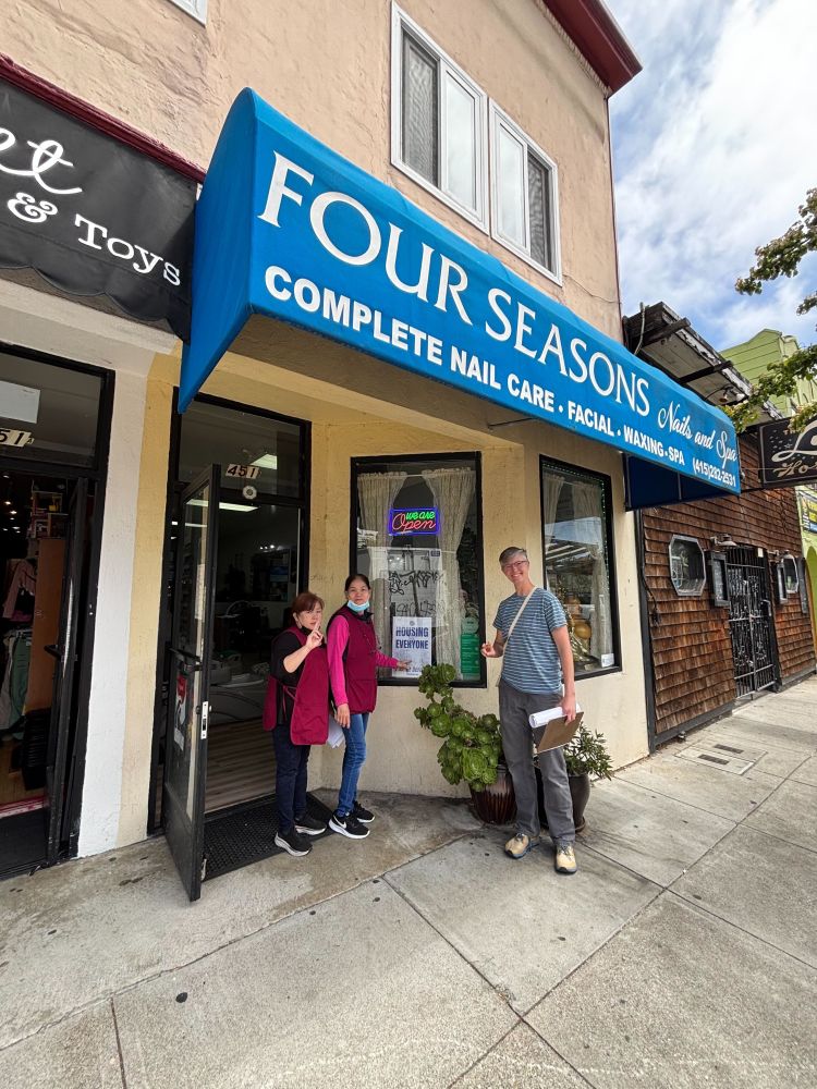 A D9Neighbors for Housing member talking about rezoning with small business owners on Cortland Avenue, who have hung up a window sign in support of rezoning Bernal in their window.