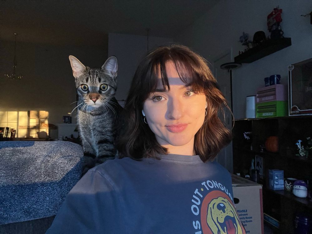 A selfie of myself and Atlas, my brown tabby cat. It’s golden hour and the back of my parent is in frame. You can see my Knick knack shelf to the right of me. My hair is brown and shoulder length. Atlas is looking just under the camera with wide eyes. He is over the left side of my shoulder. I’m wearing one of my “life is good” shirts with the phrase “Suns out • Tongues out” with a yellow lab on the front with his tongue out. 