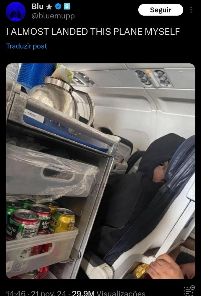 Foto de um passageiro sentado numa poltrona de  avião. Ele tá com um capuz de moletom tampando o rosto, mas com a orelha meio de fora, com um fone de ouvido pequeno, que gera uma ilusão de ótica - parece que tem alguém de capuz com o olho arregalado olhando diretamente pra quem tirou a foto