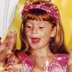 Foto em close do rosto da Marina Ruy Barbosa criança, aparentemente cantando parabéns. Ela tá com uma roupa rosa e um enfeite na cabeça parecendo uma coroa, no mesmo tom de rosa da roupa.