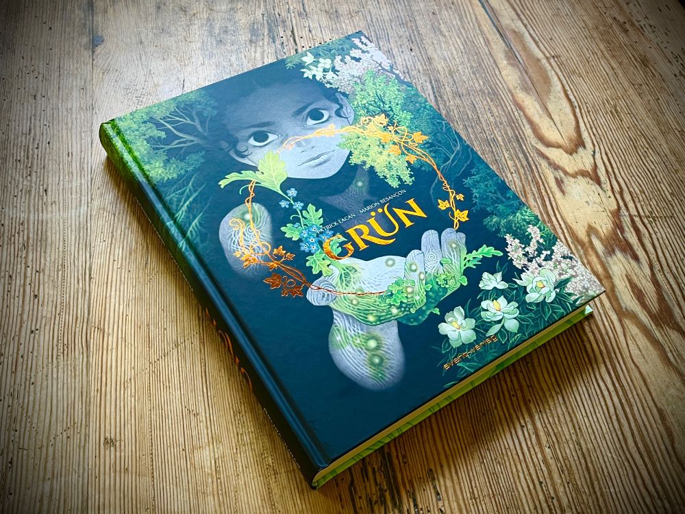 Ein Hardcover-Album (Comic) liegt auf einem markant gemasterten Holztisch. Das Cover von GRÜN zeigt ein Frauengesicht, das aus Wald/Büschen kommt, im Vordergrund eine von Grün überwucherte, mutierte, ausgestreckte Hand. Typo ist golden. (Verlag: Reprodukt)