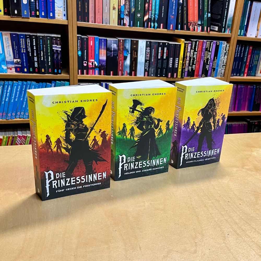 Drei Bücher stehen seitlich leicht versetzt in einer Buchhandlung auf Holztisch vor Buchregalen: Die Prinzessinnen: Fünf gegen die FInsternis, die Prinzessinnen: Helden und andere Dämonen, Die Prinzessinnen: Hoheitliches Gemetzel, von Christian Endres, Cross Cult. 