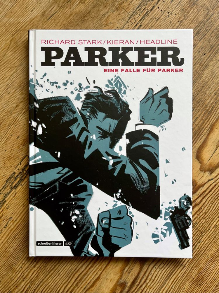 Weiß-blau-schwarzer Comic auf Holzhintergrund. Cover des Comic-Albums "Parker: Eine Falle für Parker". Ein Mann springt durch ein Fenster, verliert seine Pistole, schützt sein Gesicht mit dem Arm vor Splittern 
