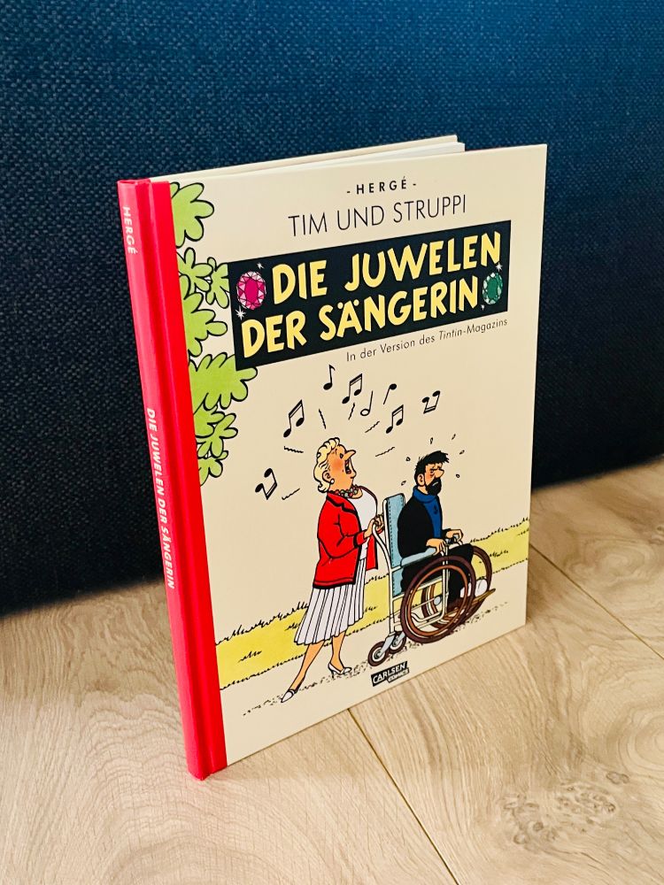 Das "Tim und Struppi"-Album "Die Juwelen der Sängerin" in einer neueren Sonderausgabe als HL-Hardcover. Aufrecht stehend auf hellen Holzboden, vor dunklem Hintergrund, leicht schräg. 