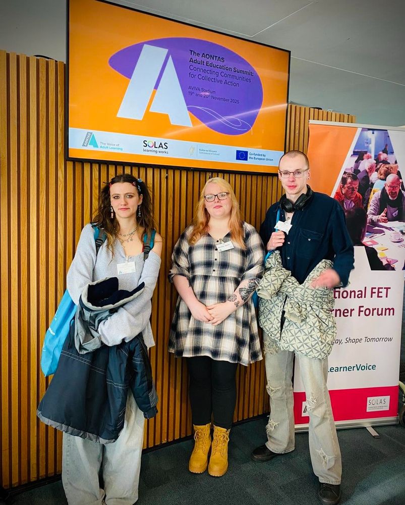 Three people at a conference. There is a branded pop-up banner and a screen behind them.