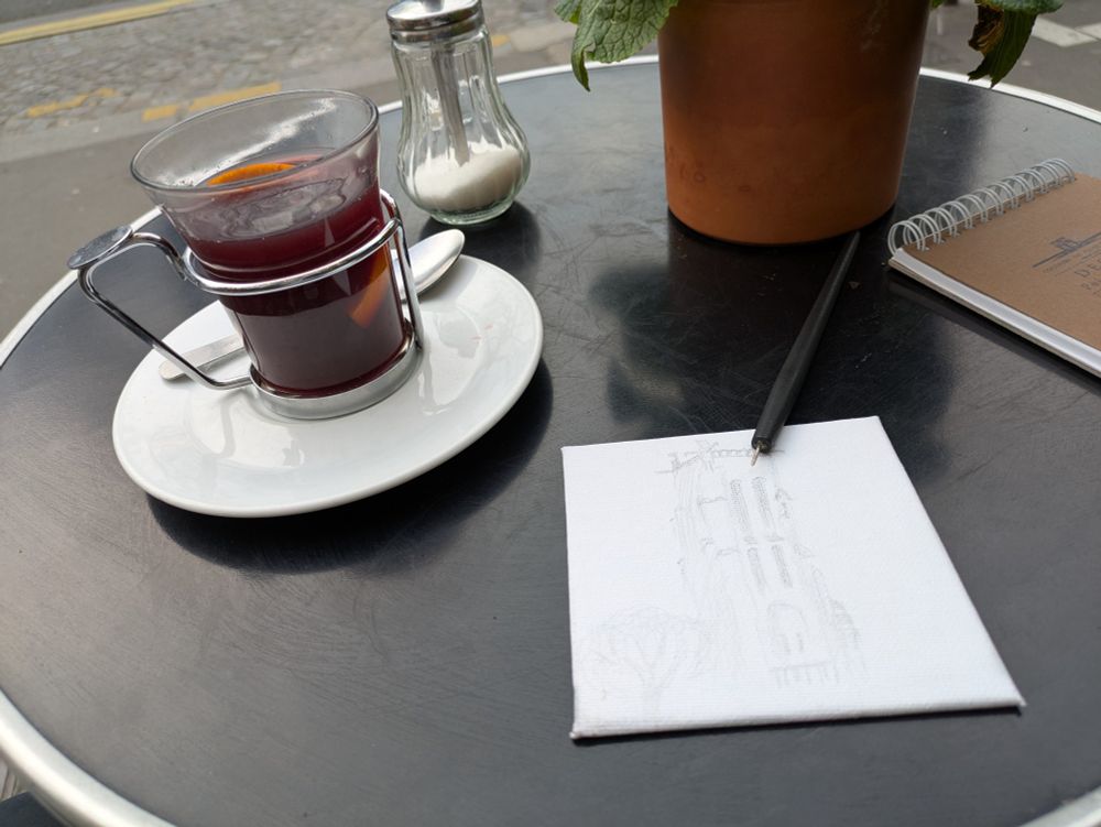 An outside table of a small bistro in Paris, with yellow flowers, a sugar shaker, a glass of mulled wine, a canvas board with a drawing, a silver stylus - a piece of fine silver wire glued to a handle - and a small sketch book.