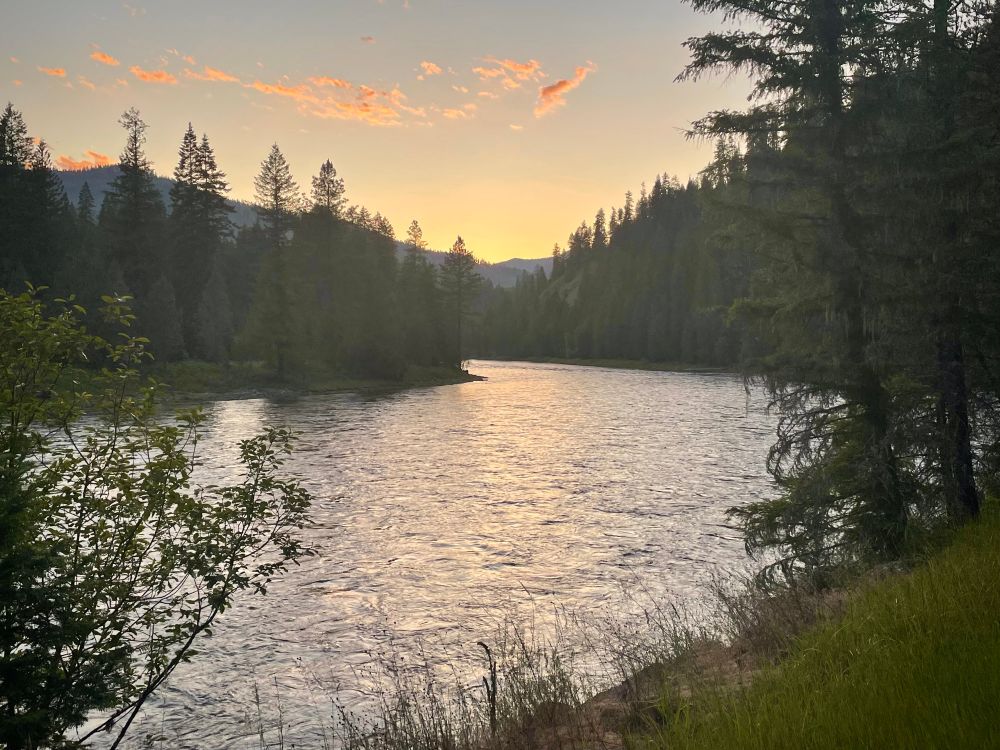 Sunset over a river cutting between forested mountains. Golden sun lights up the clouds. 