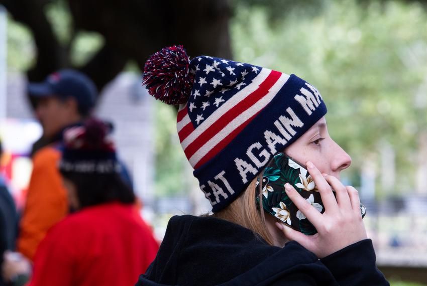 A young woman is captured in a medium shot, from the side, engrossed in a phone call. Her blonde hair is partially obscured by a dark jacket. A striking knit cap adorns her head, featuring the Stars and Stripes pattern of the American flag, complete with a pom-pom on top. The text 