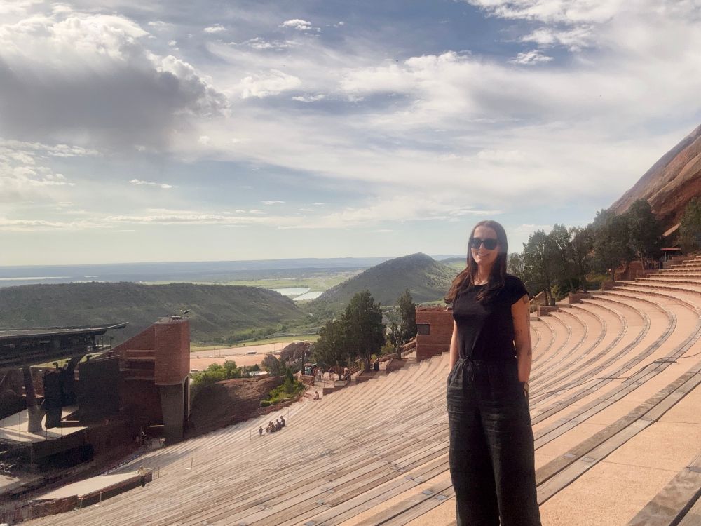 A photo of me standing on the stairs of Red Rocks Amphitheater, with light clouds in the sky and view of the green Colorado landscape in the back.
