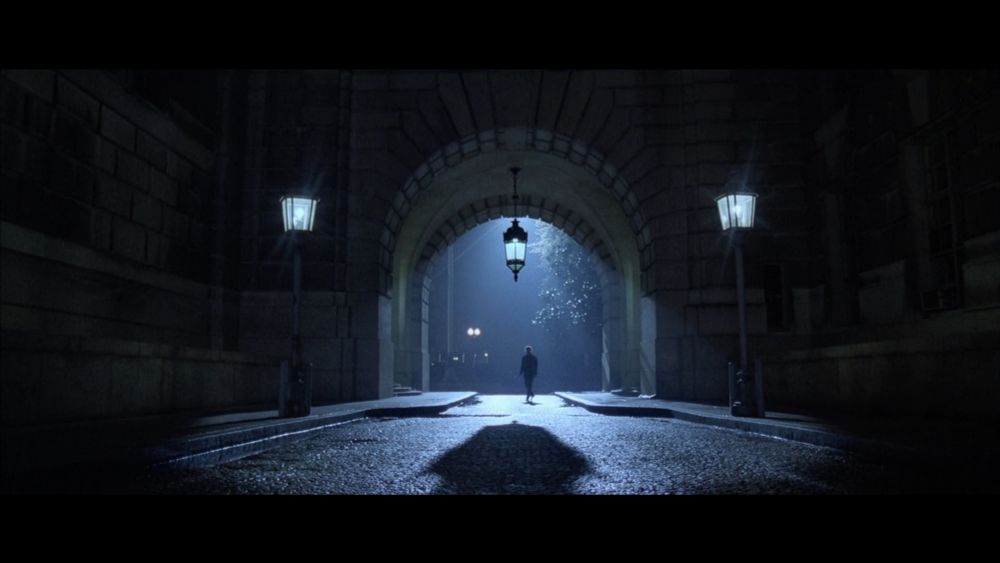 A wide arch in a large building, with old fashioned street lamps to either side, and a lamp hanging in the centre of the arch.

This creates a cone of light on a darkened street with a shadow apparently belonging to a lone figure walking towards the camera through the arch.