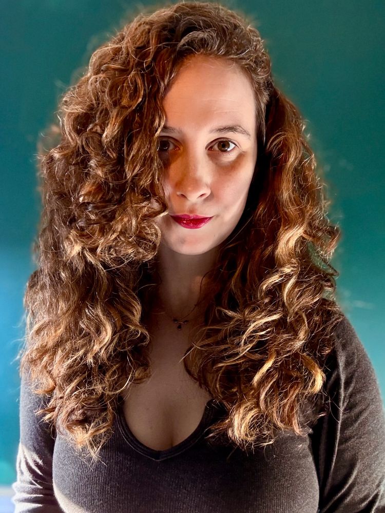 A portrait of a woman with long, curly brown hair and fair skin, standing against a teal background. She is wearing a dark V-neck top, with natural light highlighting her hair and features. She has a subtle smile.