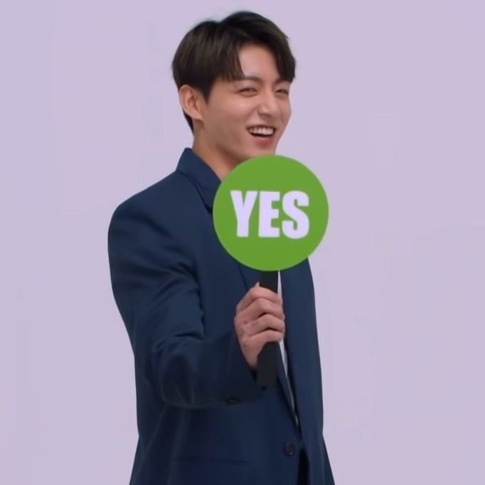 Jungkook wearing a dark blue jacket holding up a green paddle that reads "YES" and making a winky face
