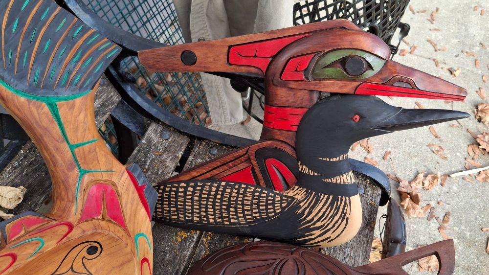 A beautiful wood carving by the artist Kevin Touchie, in yellow cedar, rests on a wood and metal bench. The carving shows a realistic loon transforming into a spirit bird that is done in the style of the Nuu-Chah-Nulth tribe on the Salish coast of BC.