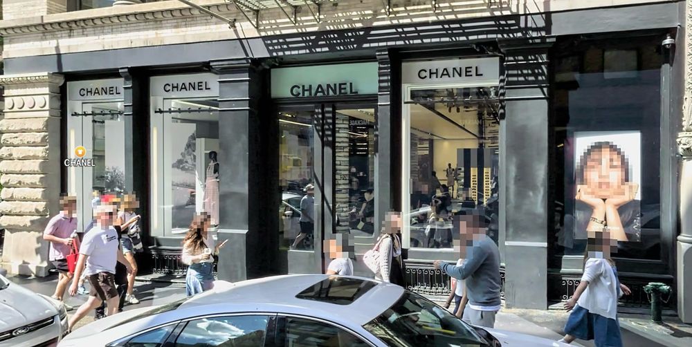 A sidewalk and Chanel store in Manhattan, in a photo taken firm a passing car. About ten people are on the sidewalk, and there faces were blurred out for anonymity. There's a large ad in the store window, with a picture of a woman facing out to the street, and her face is also blurred out.