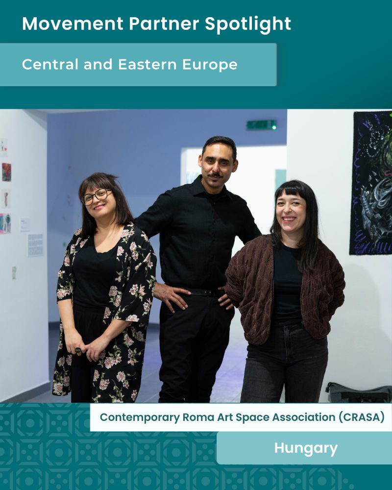 Three people stand side by side in a contemporary art gallery space, smiling warmly at the camera. On the left is a woman with long dark hair, wearing glasses, a black top, and a black floral-patterned kimono-style jacket. In the center is a man with short dark hair and a mustache, wearing a black shirt tucked into black trousers, with one hand resting on his hip. On the right is a woman with long dark hair and bangs, wearing a dark brown textured jacket and dark jeans. Behind them, the gallery walls display colorful artwork and informational posters. The image is framed with a teal graphic design overlay. At the top, bold text reads “Movement Partner Spotlight” and “Central and Eastern Europe.” At the bottom, a text box identifies the group as “Contemporary Roma Art Space Association (CRASA)” and notes their location as “Hungary.”