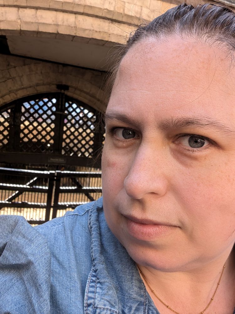 Image is a selfie, taken by Amy. She is wearing a denim dress. Behind her is Traitors Gate, which used to allow boats from the River Thames into the Tower of London.