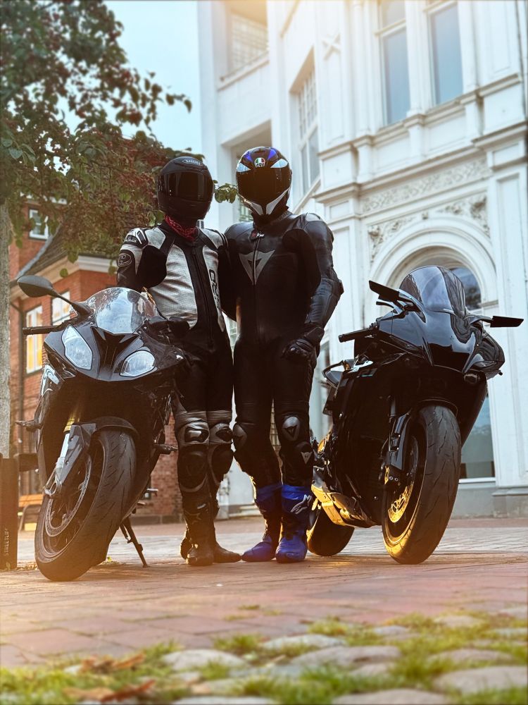 Two motorcycle riders with their bikes in the sunset in front of old houses.
