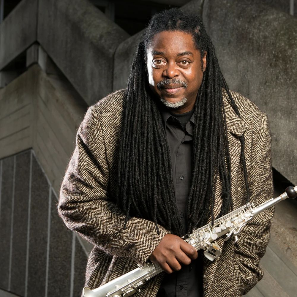 Courtney wearing a tweed jacket and black shirt smiles holding a silver alto saxophone next to a brutalist background