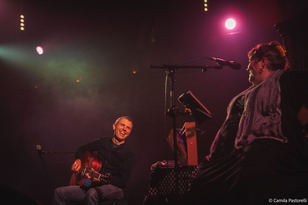 The Breath laughing and looking at each other sitting on stage with their instruments and microphones