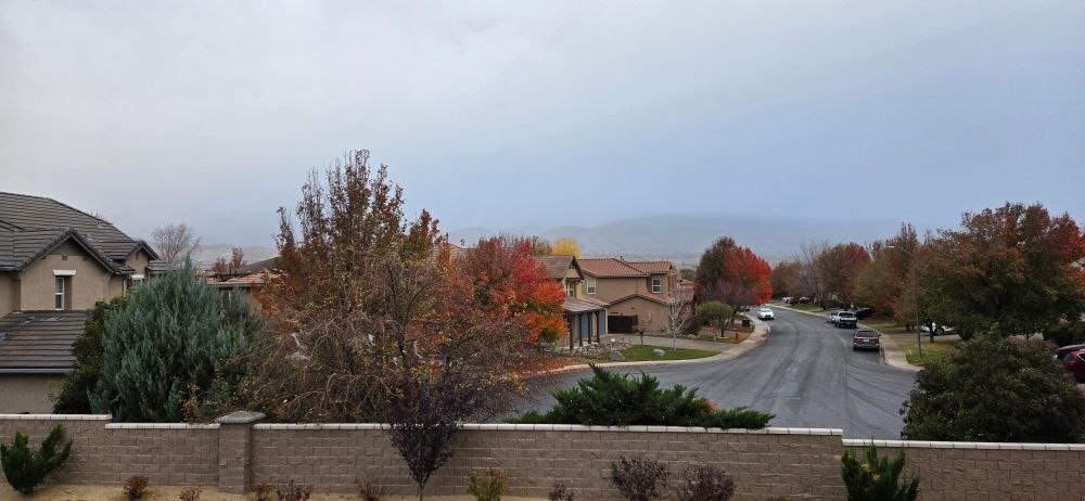 Colorful autumn trees flank a damp street where tire tracks are visible in the gathering moisture. Looking west from the Syeamboat/Diamonte Ranch area of south Reno, rain clouds, shroud the mountains that are usually visible.