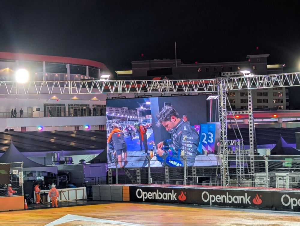 somewhat zoomed in shot of the same screen showing f1 racer carlos sainz in williams blue with fluffy messy hair getting ready to put on a ballcap