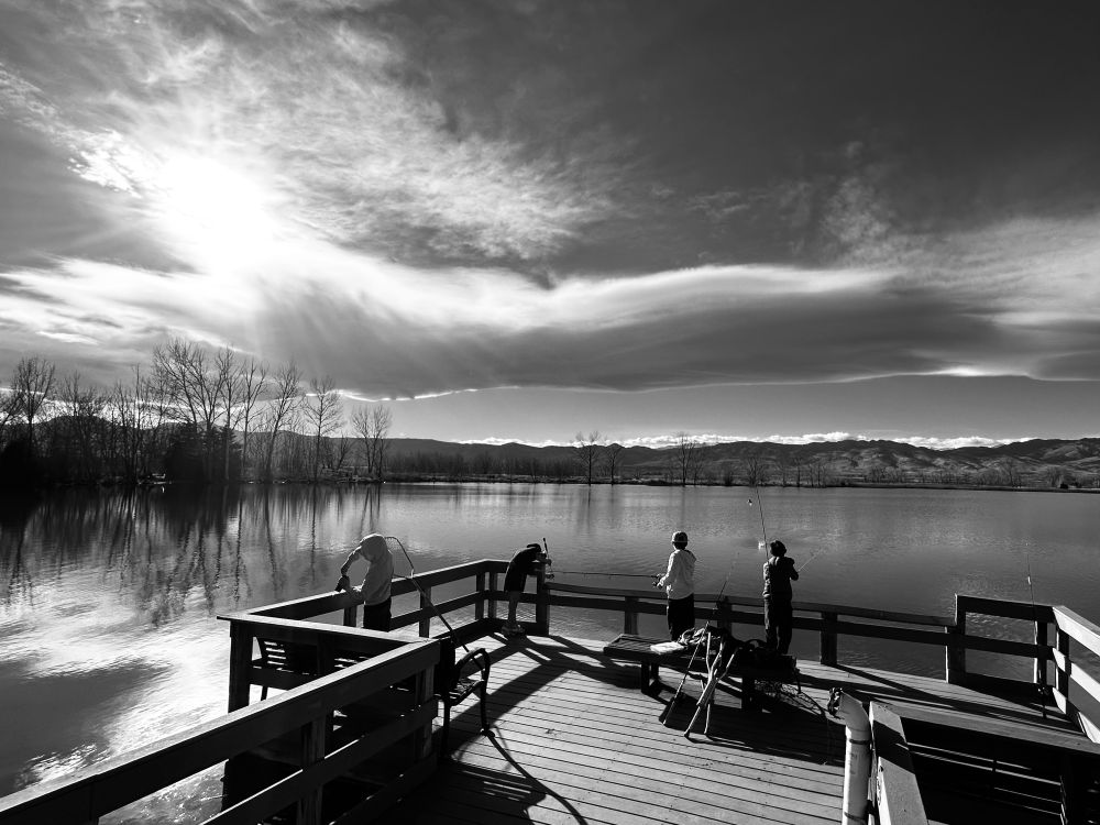 Four children on a dock fishing.   Clouds and the setting sun in the sky and reflected on the water.   The foothills appear to surround the lake.