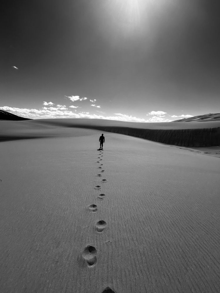 A man walks towards the sun across a giant sand dune leaving a solo set of footprints behind. 