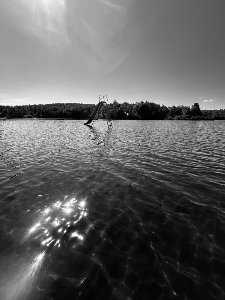 A slide sits out from shore in a lake.  The forest forms a boundary behind it.  Rays from a full sun shine in the sky and our reflected in the water closer to shore.