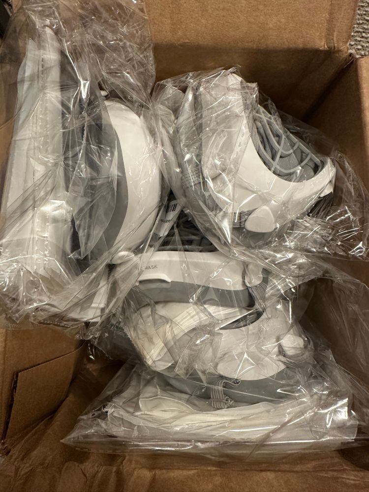 Brown cardboard box full of white and grey Flo Mask respirators and filters piled on top of each other. 

