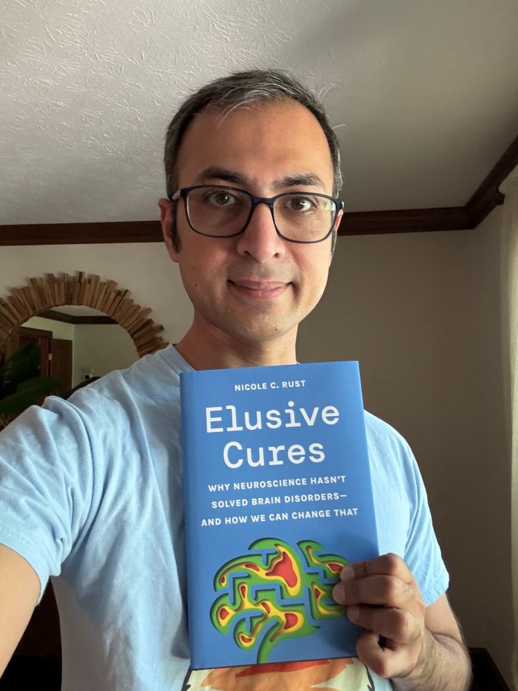 A man wearing glasses and a light blue T-shirt is standing indoors, holding a book titled Elusive Cures by Nicole C. Rust. The book’s cover is bright blue with a colorful abstract illustration of a brain at the bottom. The subtitle reads: “Why neuroscience hasn’t solved brain disorders — and how we can change that.” The background features a beige wall, a wooden-framed circular mirror, and a window with cream-colored curtains. The lighting is natural and soft.