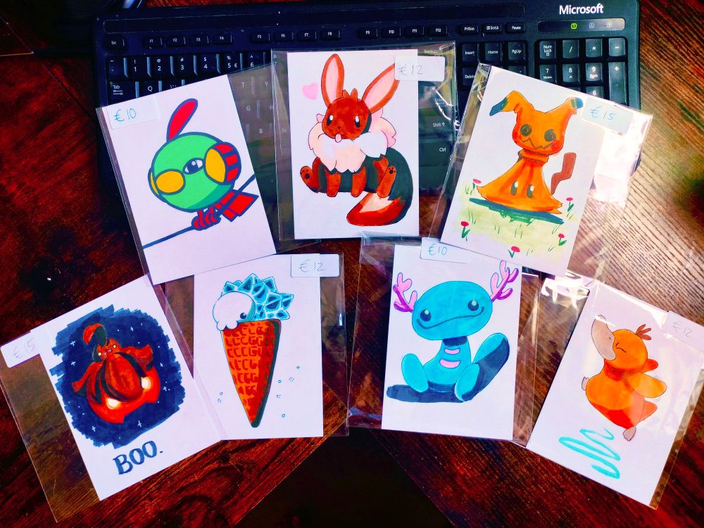Seven drawings on a desk. One posca natu, and six marker drawings of eevee, mimikyu, pumpkaboo, snom on an icecream cone, wooper and psyduck