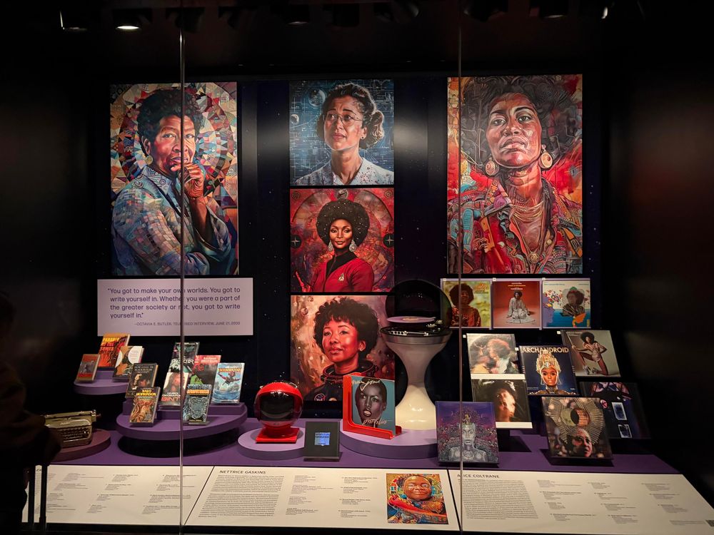 A display case filled with artwork and memorabilia celebrating black women in science fiction, including books, vinyl albums, a retro futuristic space helmet, and hyper color portraits. Center stage in Nichelle Nichols as Utica from Star Trek.