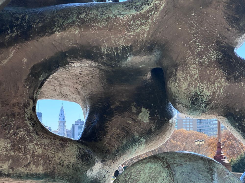 A detail of a sinuous metal sculpture by Jacques Lipchitz. There is an aperture through which you can see a blurry version of Philadelphia City Hall in the distance.
