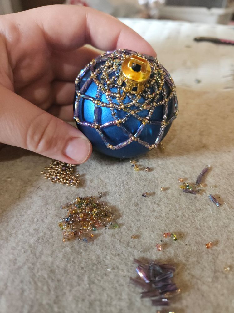A blue Christmas ornament with a beaded over netting in tones of cold, copper, blues and purples. Beads are nearby.  