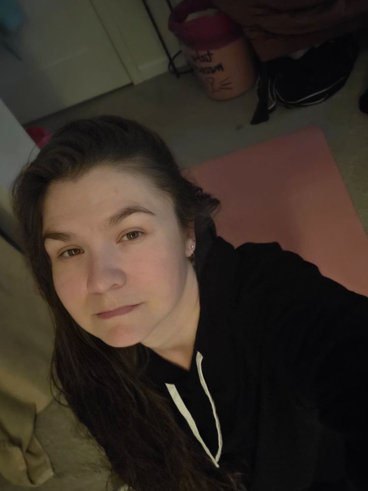 An unimpressed and pasty human in a black sweater sits on a pink yoga mat. They stare vaguely at the camera. While there technically is a reflection of light in their eyes from a nearby lamp, a discerning person might be able to tell that this human is completely over the concept of participating in capitalism.