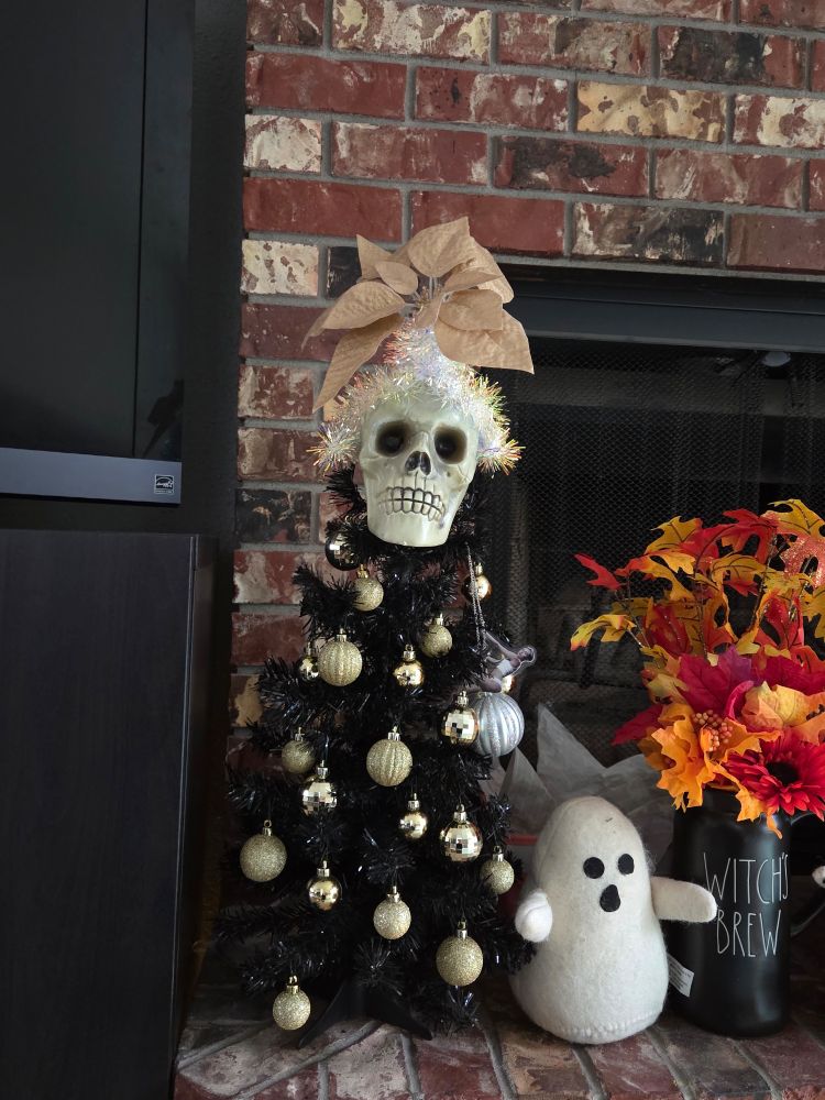 A small black plastic tree is hung with golden baubles, and the tree topper is a plastic skull. The skull has a little glittery crown, the center of which is a golden poinsettia. 