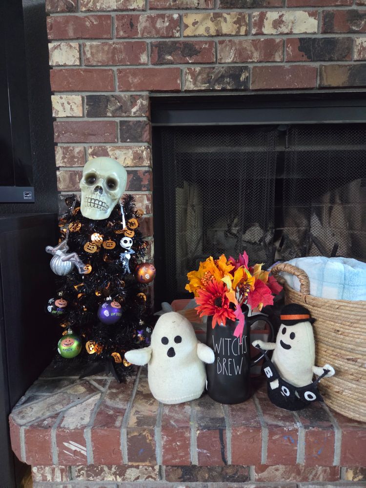 A small black plastic tree sits on a fireplace hearth. It is decorated with holiday orbs featuring pumpkins and ghosts in the colors of green, yellow, black, and orange. The usual skull is in place for the tree topper, and the Miley Cyrus wrecking ball ornament is in its honorable place. 
Next to the small tree is a little stuffed ghost, a black flower pitched labeled WITCH'S BREW filled with fake autumnal foliage, and another small stuffed ghost wearing a black hat and holding a banner that reads, "BOO".