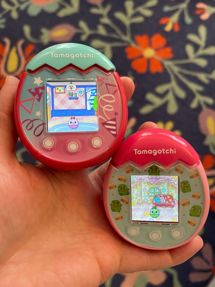 Two Tamagotchi Pix in my hand. One is pink and turquoise and the other is mostly pink with a green Kuchipatchi and burgers faceplate. 
